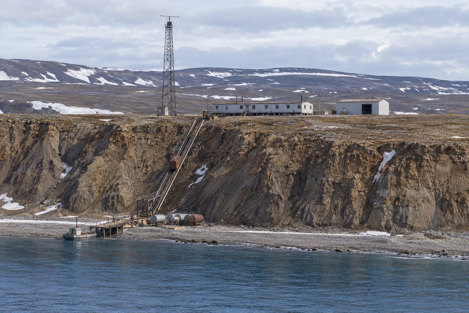Interpretive view of a small DEW Line intermediate station on cliffs above the Horton River coast, with a tall Doppler antenna tower, low module train, shore fuel tanks and an inclined cargo route