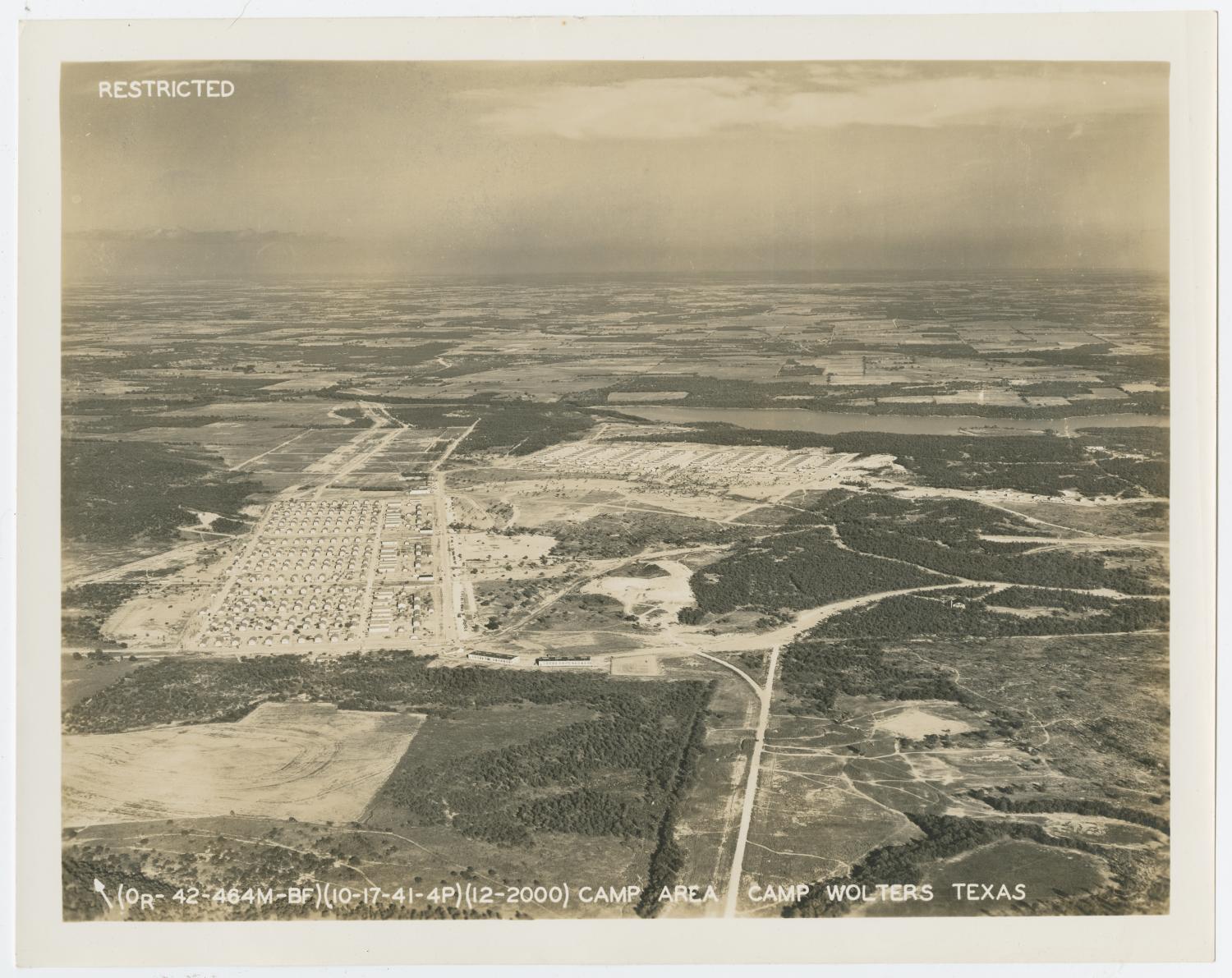 Photograph of the camp area of Camp Wolters as seen from the sky. Forests, fields, and roads surround the camp area, which is situated near the center of the photo. Rows of buildings can be seen in a section to the left and in another section closer to a body of water.