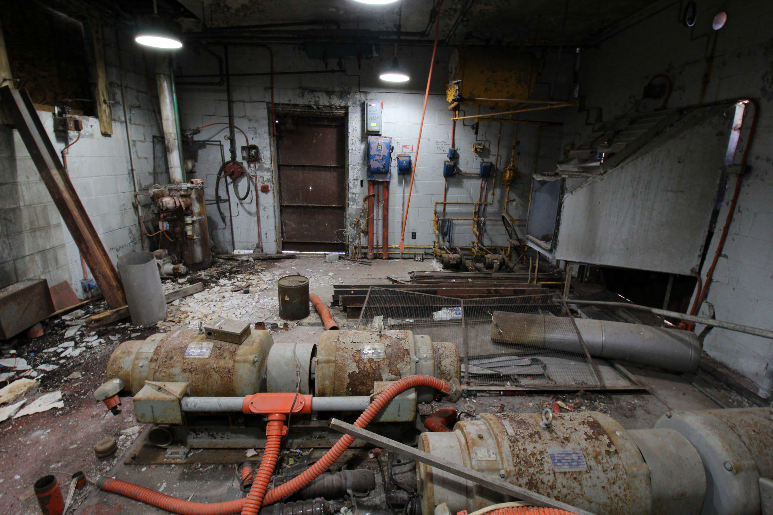 Generator room, foreground to background from west; rotary regulators, diesel generator cooling air intake, fuel pumps beyond on floor, steel plate filling door opening, diesel fired water heater left corner