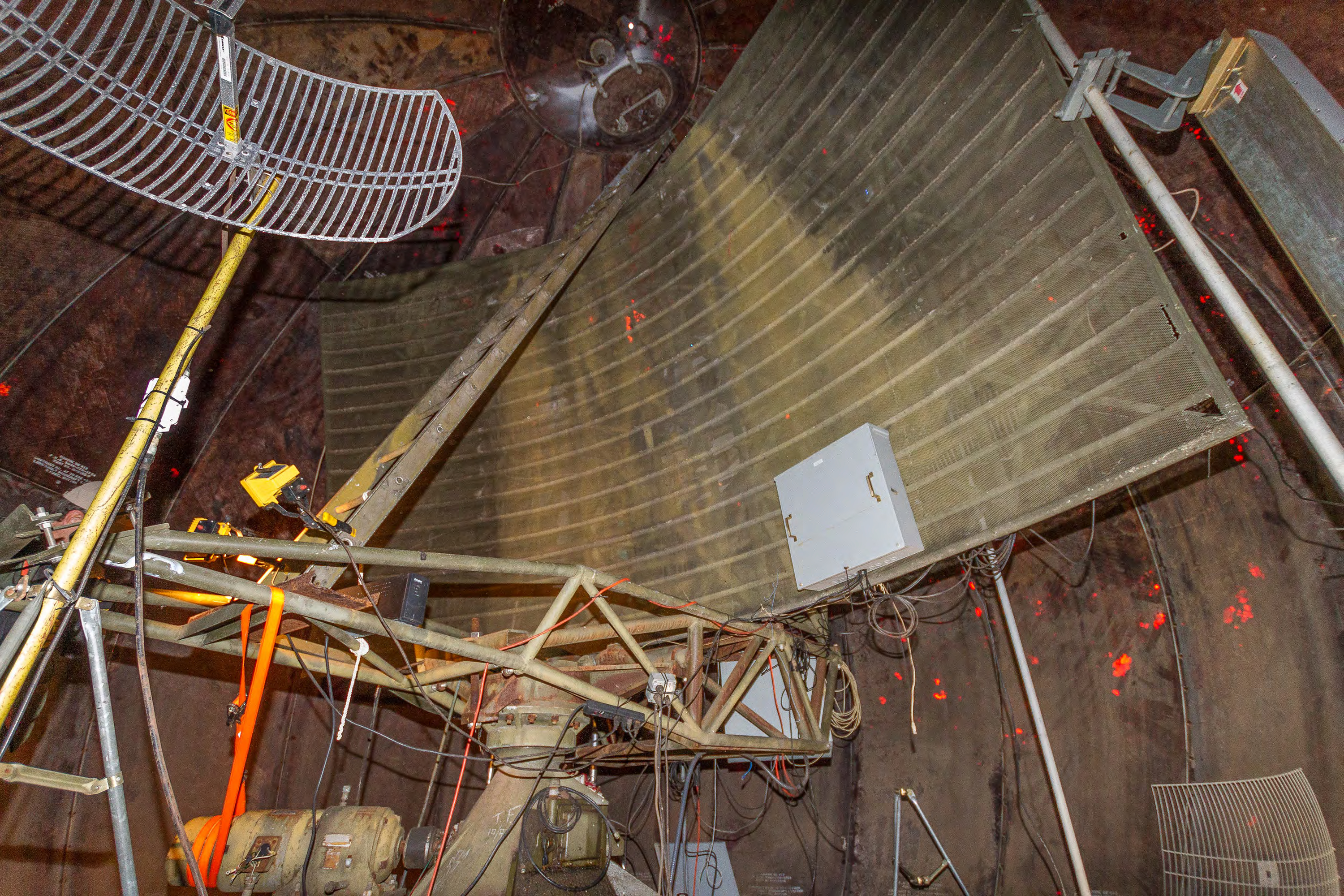 Interior of radome from door on south quadrant of antenna deck: relatively modern cellular phone and microwave antennas foreground; FPS-18 radar antenna reflector, frame, rotating pedestal, and drive motor beyond with non-original ladder to hatch; fiberglass radome panels background, sunlight filtering through areas of missing exterior paint